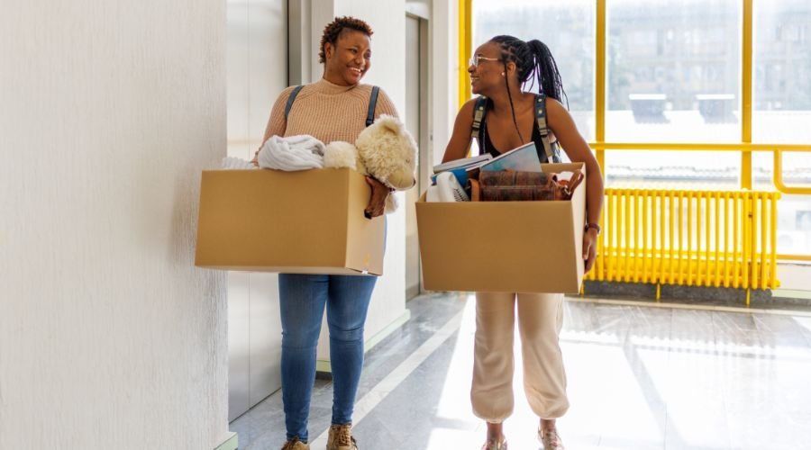 Two women carrying boxes in hallway.
