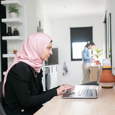 Woman using laptop, another in background cooking