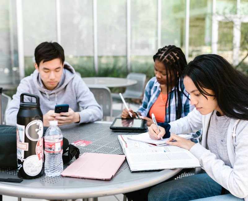 Students studying and using devices at outdoor table.